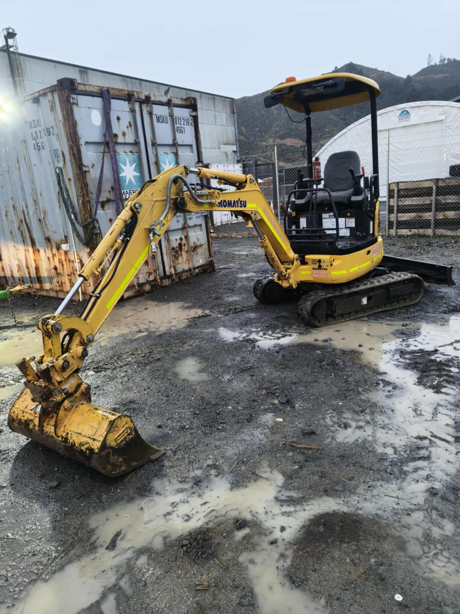 A small yellow excavator is parked in a muddy parking lot.