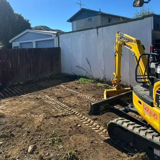 A yellow excavator is sitting in the dirt in front of a house.
