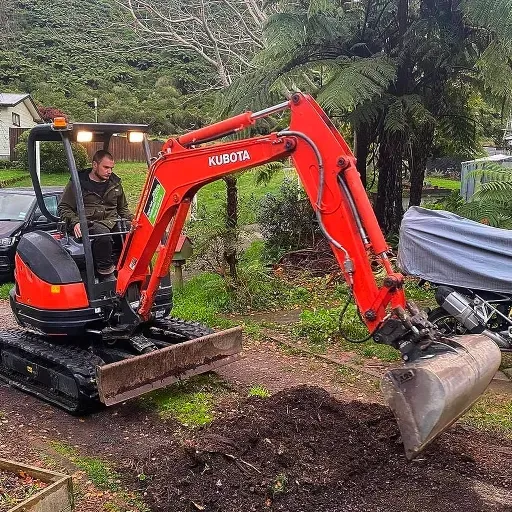 A man is driving a small red excavator in a yard.