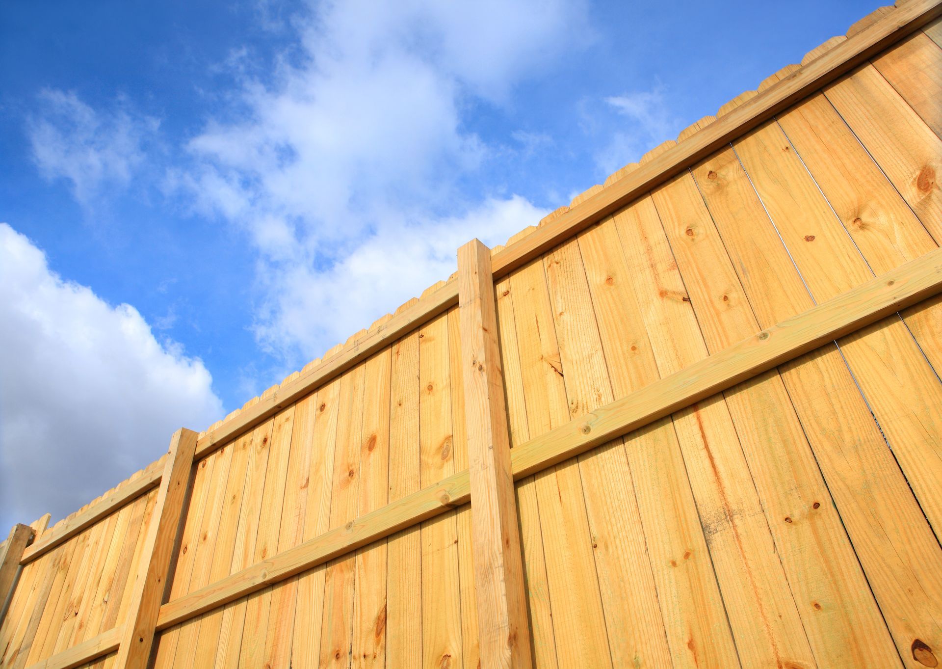 Tall wooden fence panels against a bright blue sky with scattered clouds.