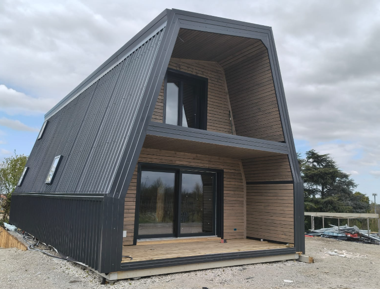 Una mini casa a due piani con tetto a capanna, rivestimento esterno in lamiera ondulata nera e pannelli in legno chiaro, situata su un lotto esterno di ghiaia.