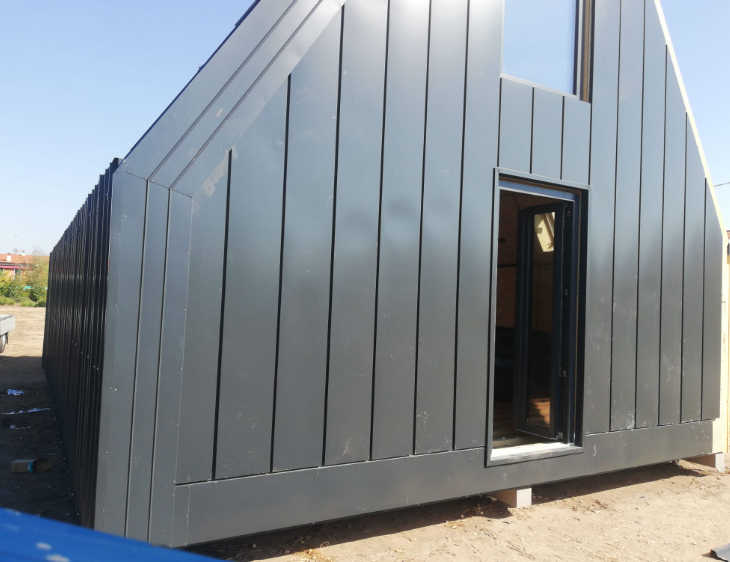 Una moderna casa modulare a forma di A con rivestimento esterno in metallo grigio a giunzione verticale e porta a vetri, situata su un lotto sabbioso.