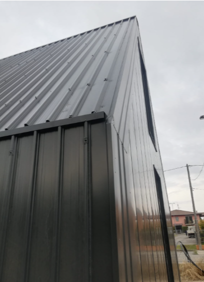 Vista dal basso di un edificio moderno con rivestimento esterno in lamiera ondulata scura e verticale e tetto a falde, sotto un cielo nuvoloso.