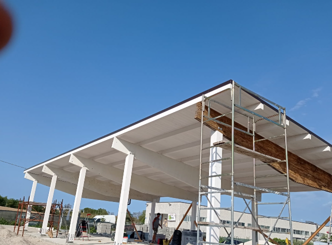 Operai edili su un'impalcatura sotto una struttura di copertura bianca ancora incompiuta, sullo sfondo di un cielo azzurro.