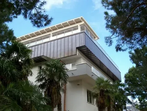 Moderno edificio a più piani con un ultimo piano metallico e un balcone, circondato da pini e palme sotto un cielo azzurro.