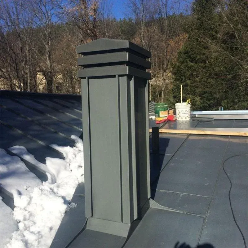Un moderno comignolo rettangolare in metallo grigio installato su un tetto in metallo scuro, con neve sul tetto e alberi sullo sfondo.