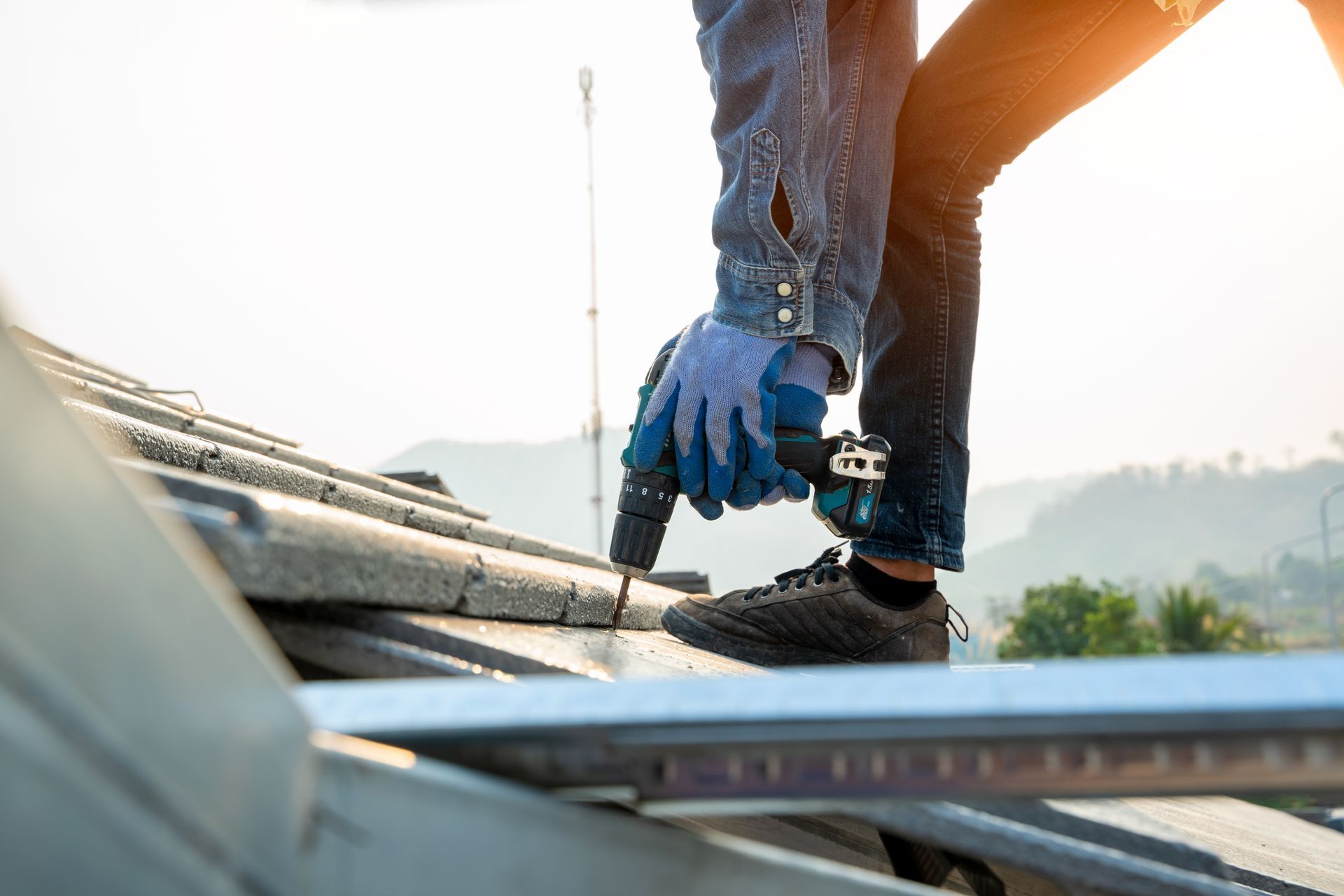 Roofer in denim shirt and gloves, using a drill to secure roof tiles on a sunny day.