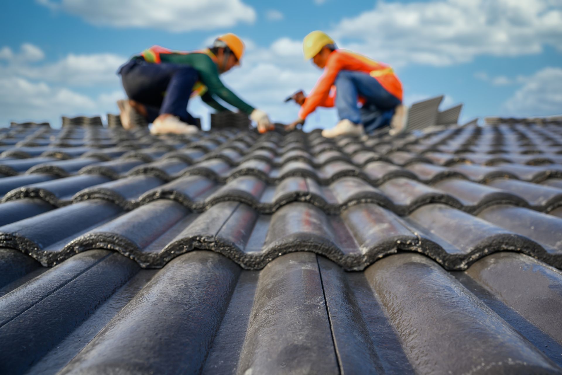 Two construction workers on a roof installing tiles; blue sky background.