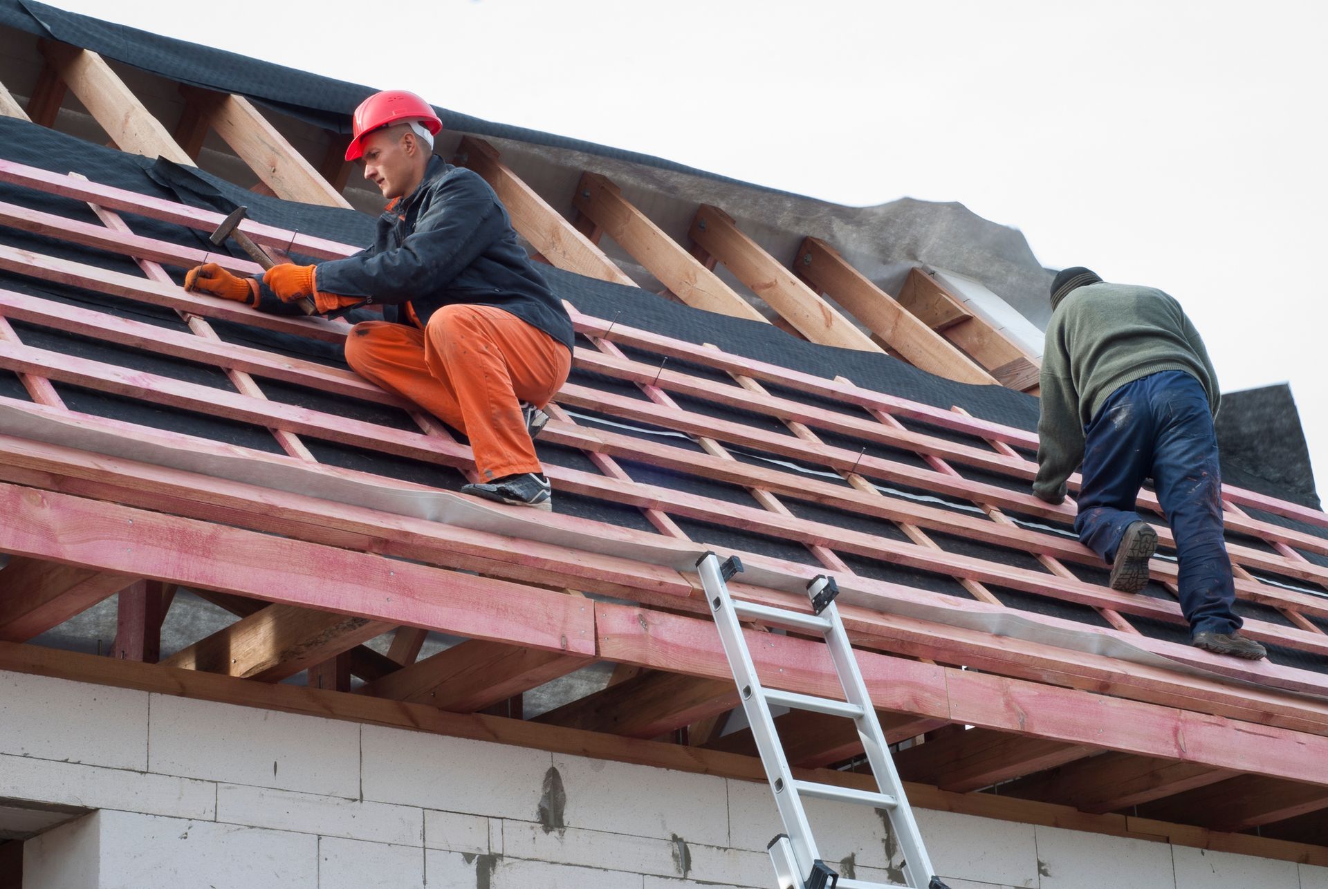 Construction workers installing roofing materials on a building frame; one uses a tool.