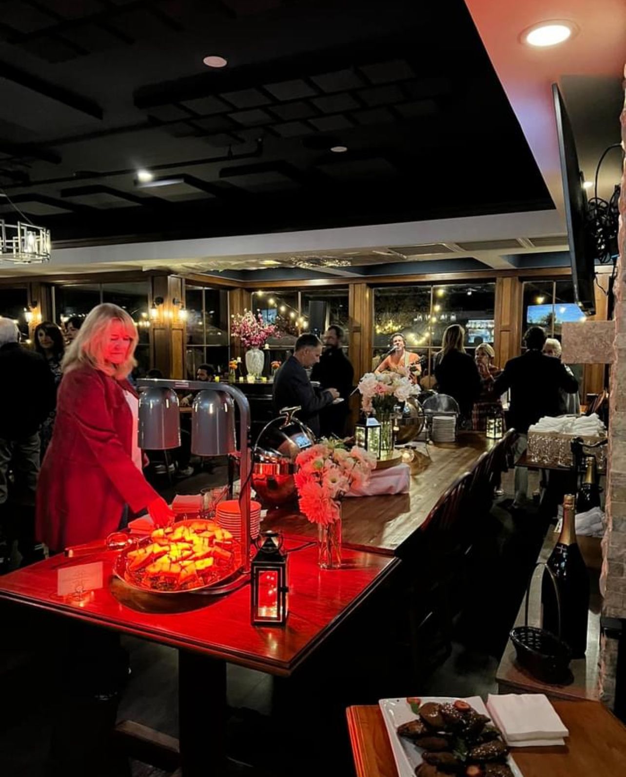 Woman at a bar in a restaurant with food; people and flowers in the background; red bar.