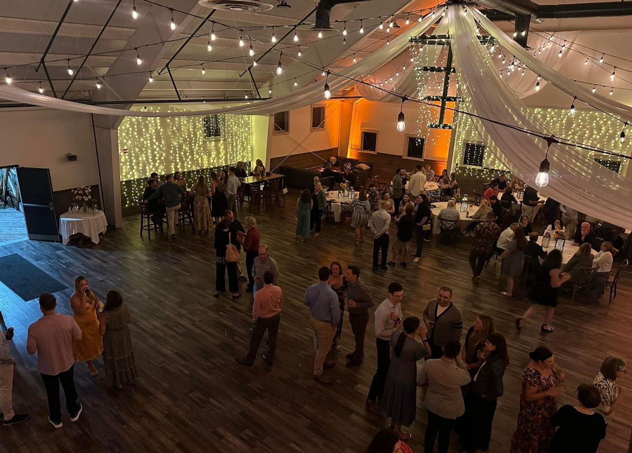 People gathering in a large room with string lights, tables, and draped fabric.