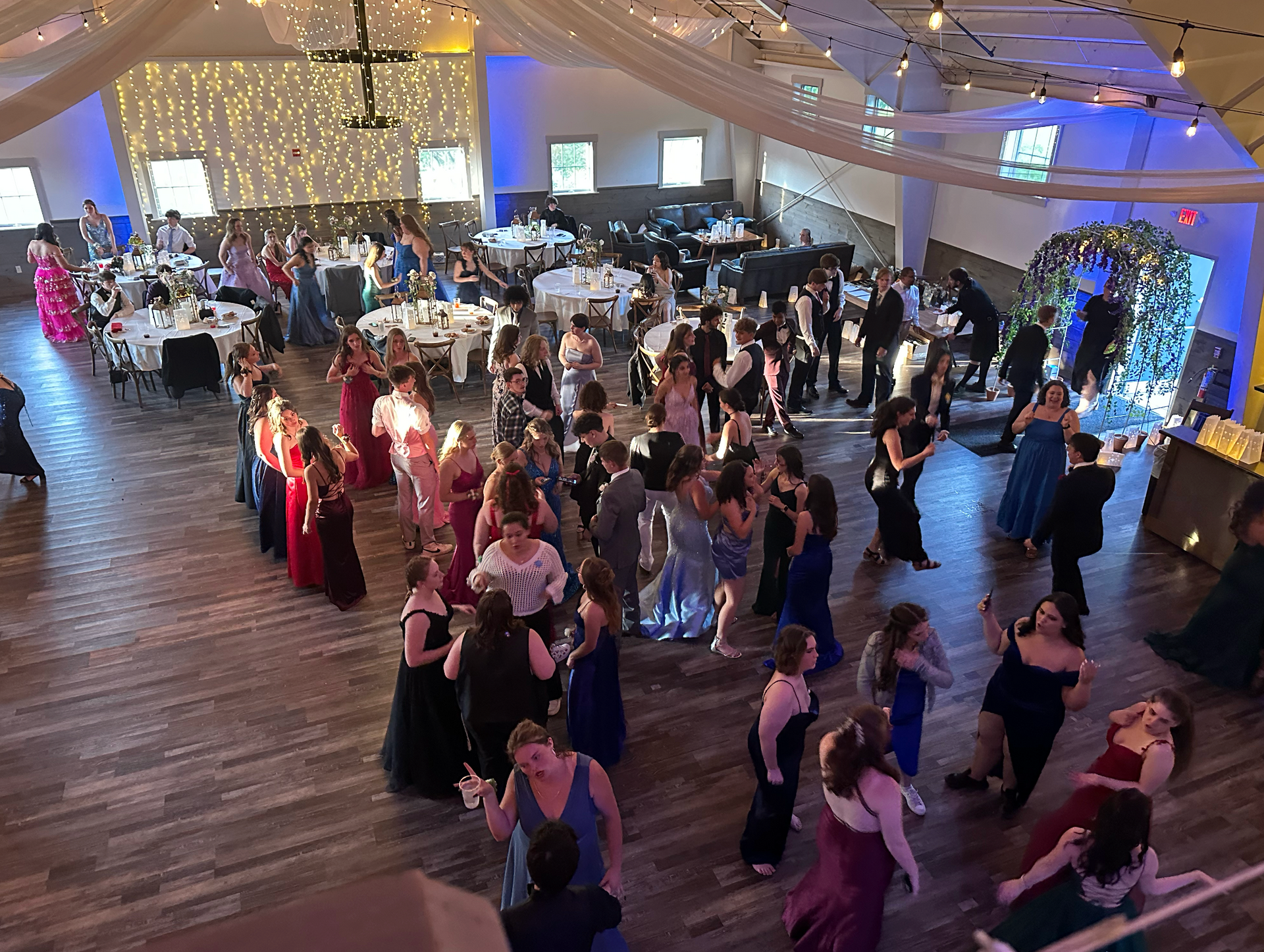 A crowded ballroom with people dancing, wearing formal attire. Tables are set with food, decorations, and lighting.