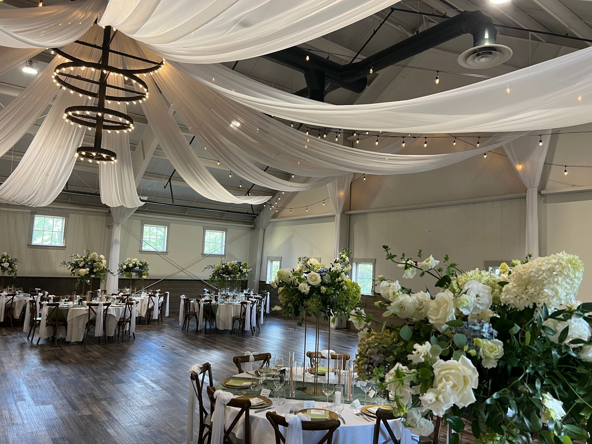 Wedding reception hall with draped white fabric ceiling, tables with floral centerpieces, and dark wooden floor.