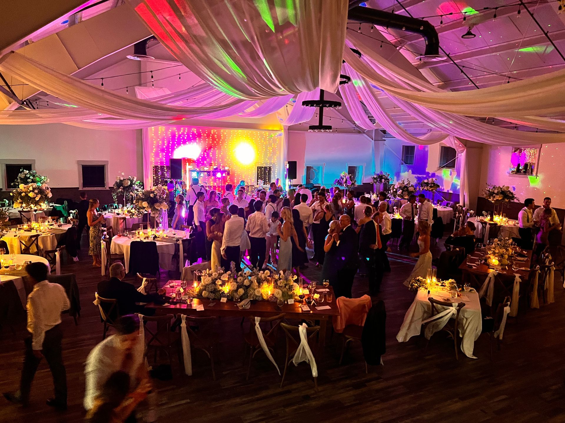 Wedding reception with guests dancing, tables set, and colorful lighting in a large hall.