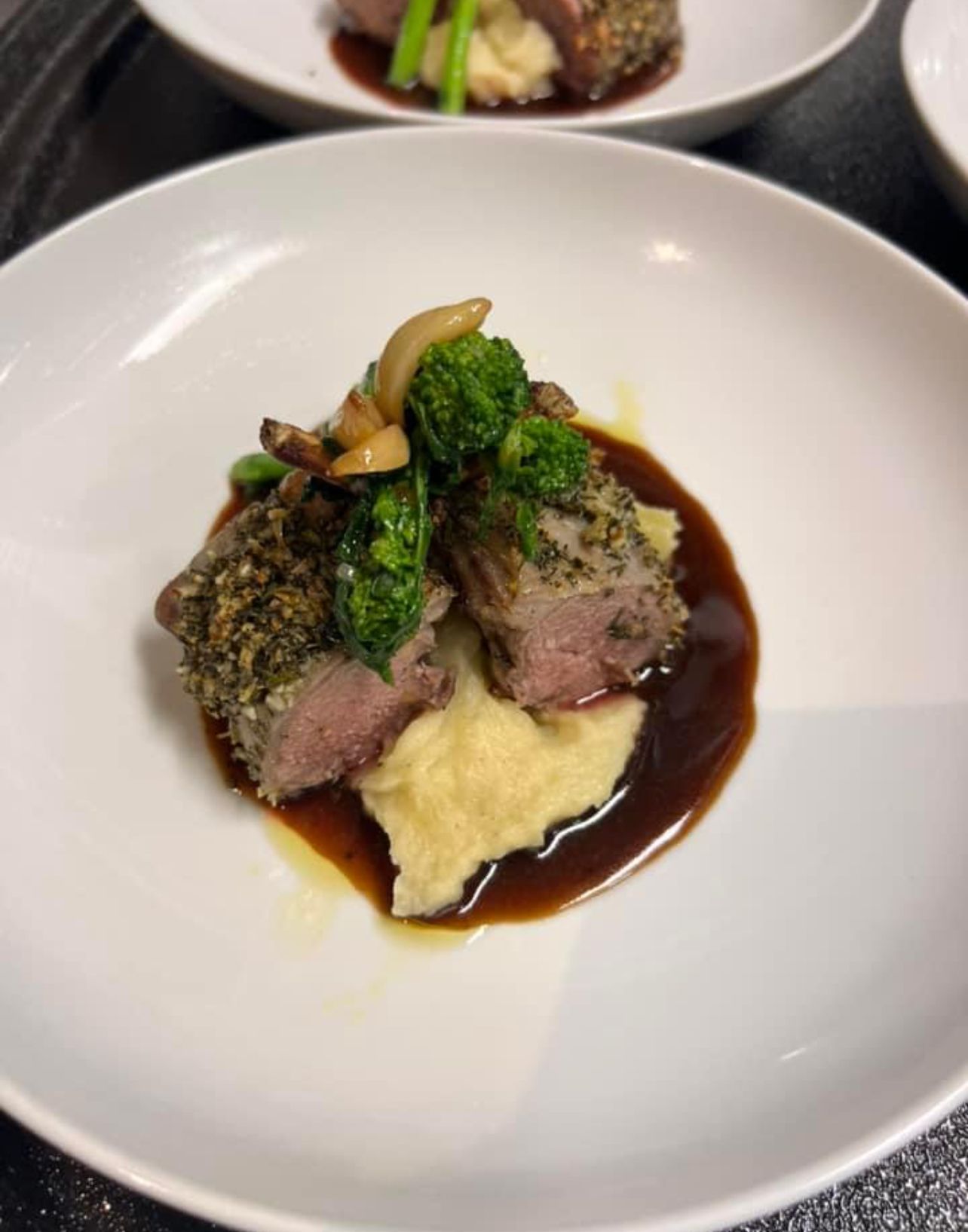 Lamb cutlet on mashed potatoes, with broccoli and mushroom in a brown sauce, on a white plate.