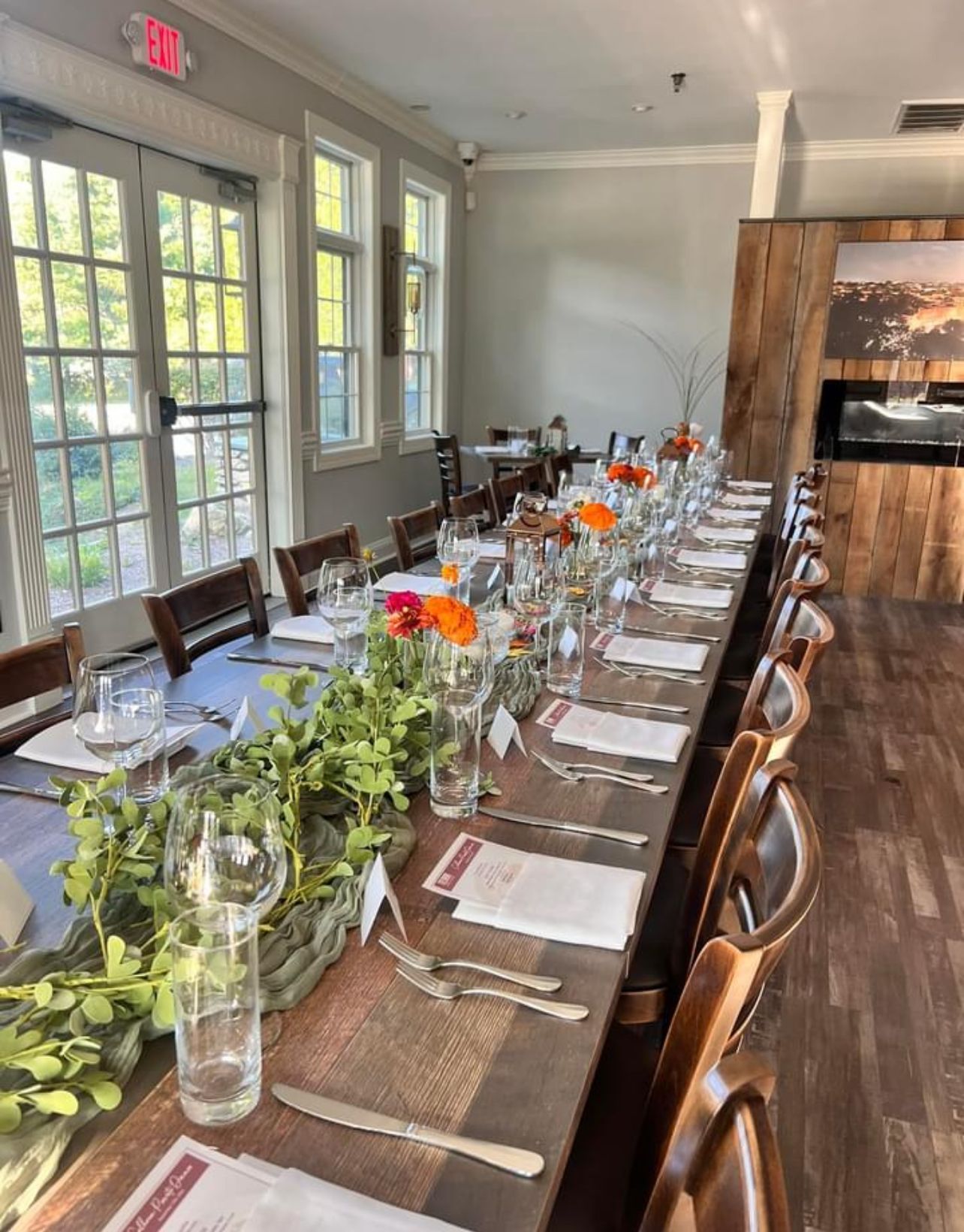 Long, decorated dining table set for a meal in a restaurant; flowers and greenery adorn the table.