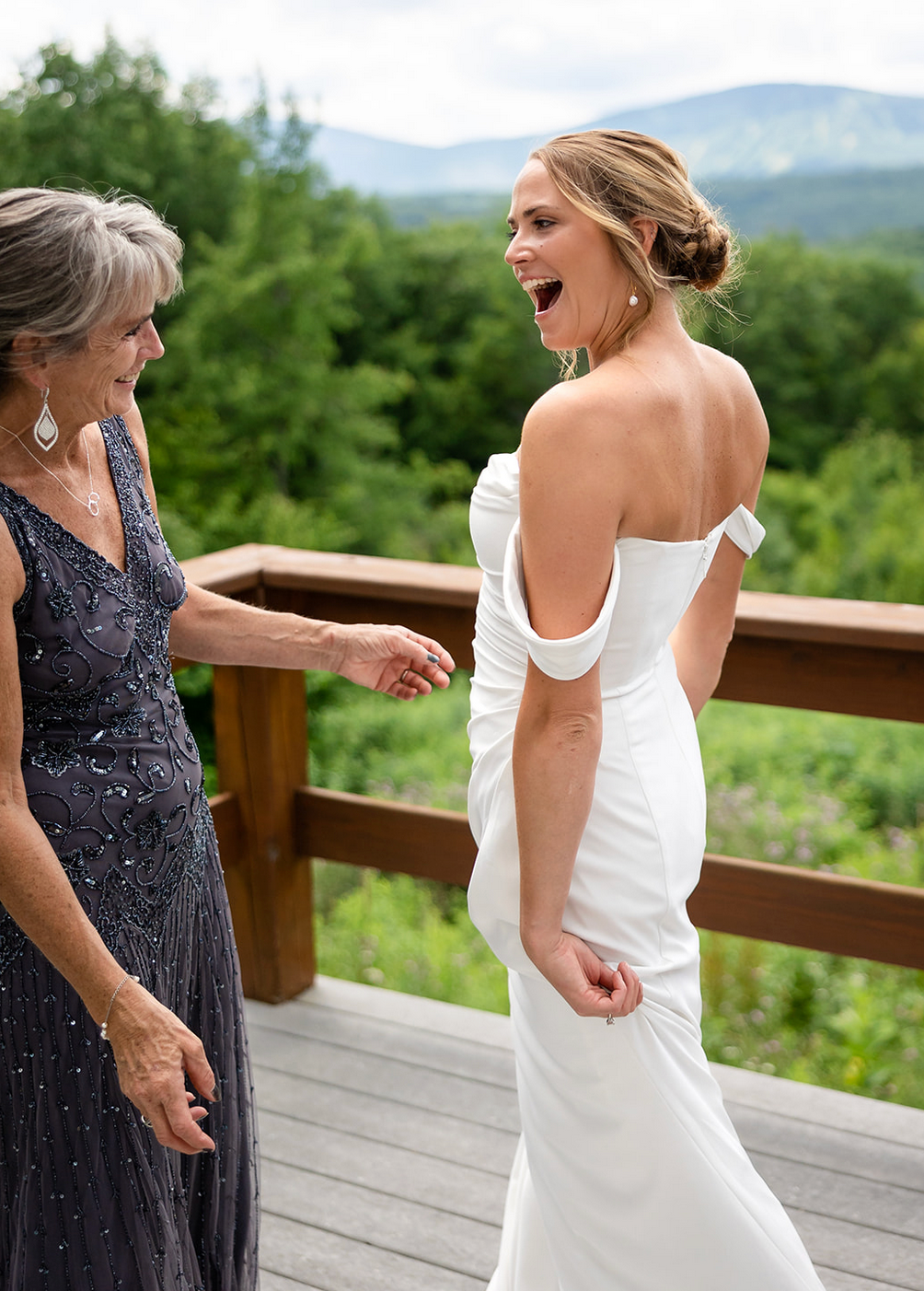 Bride in white dress laughs with a woman in a grey dress on a wooden deck overlooking mountains.
