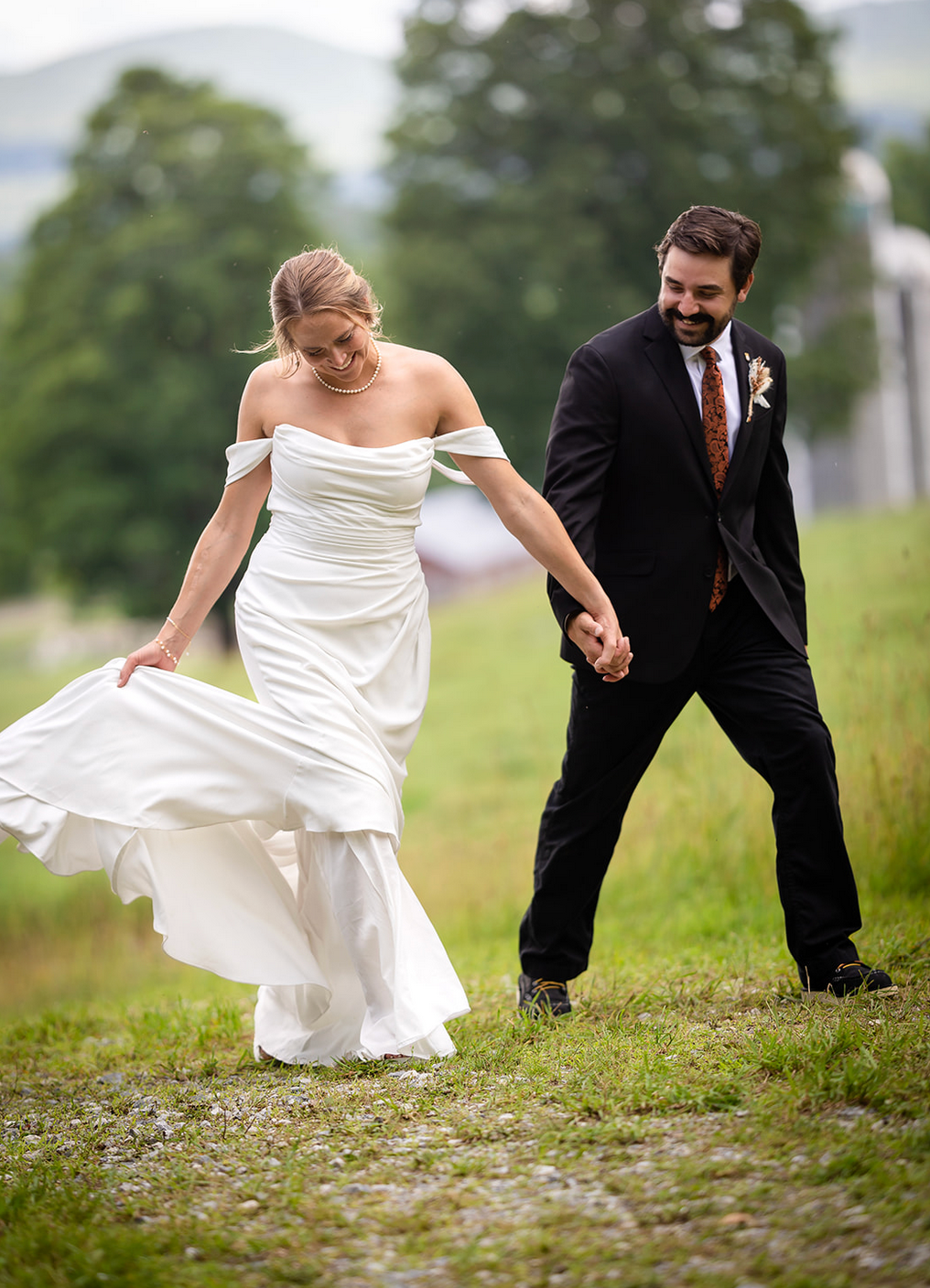 Bride in white dress and groom in suit hold hands, walking on a grassy hill.