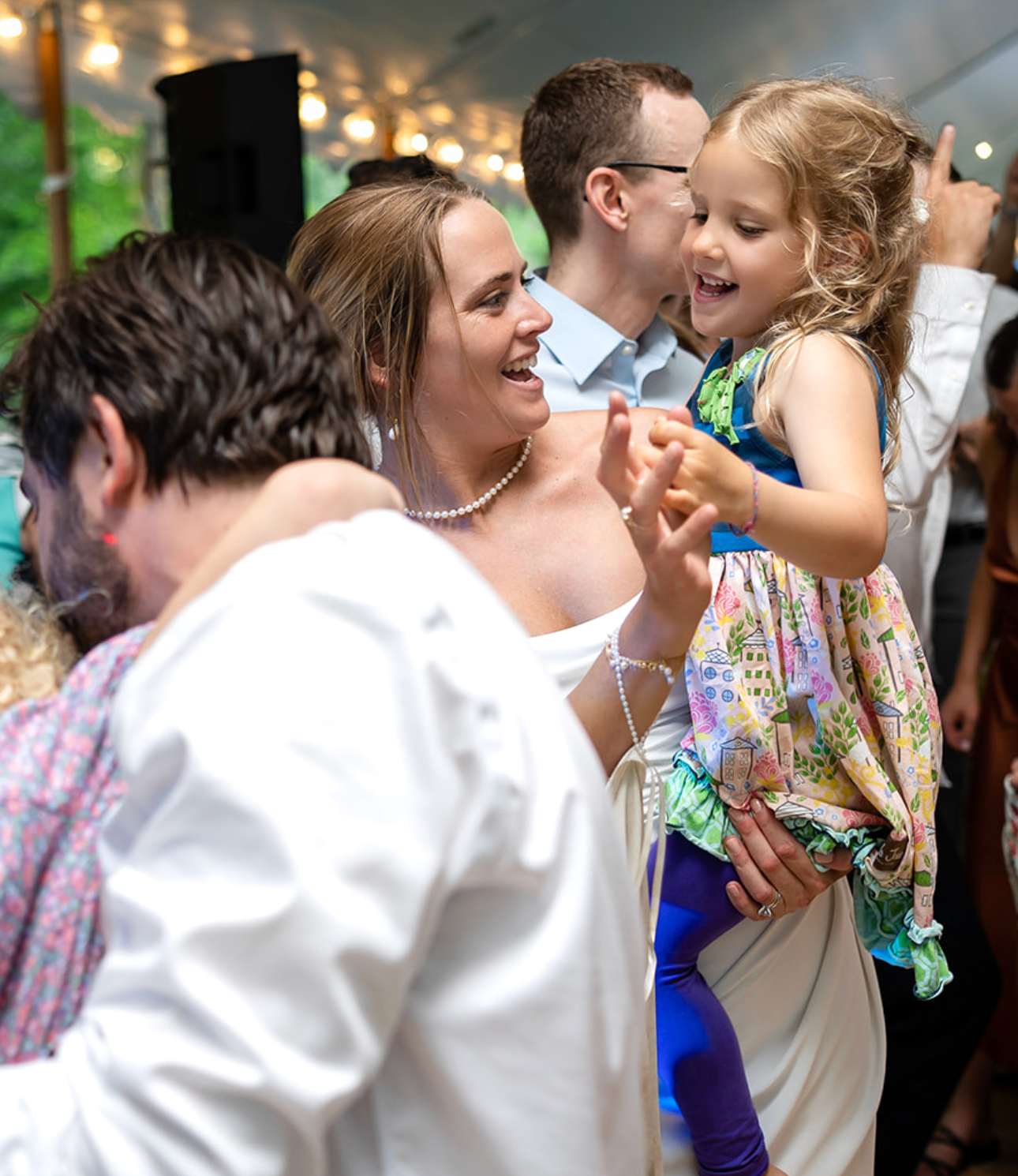 Bride dances with a child at a wedding reception; both smile brightly. People dance around them.