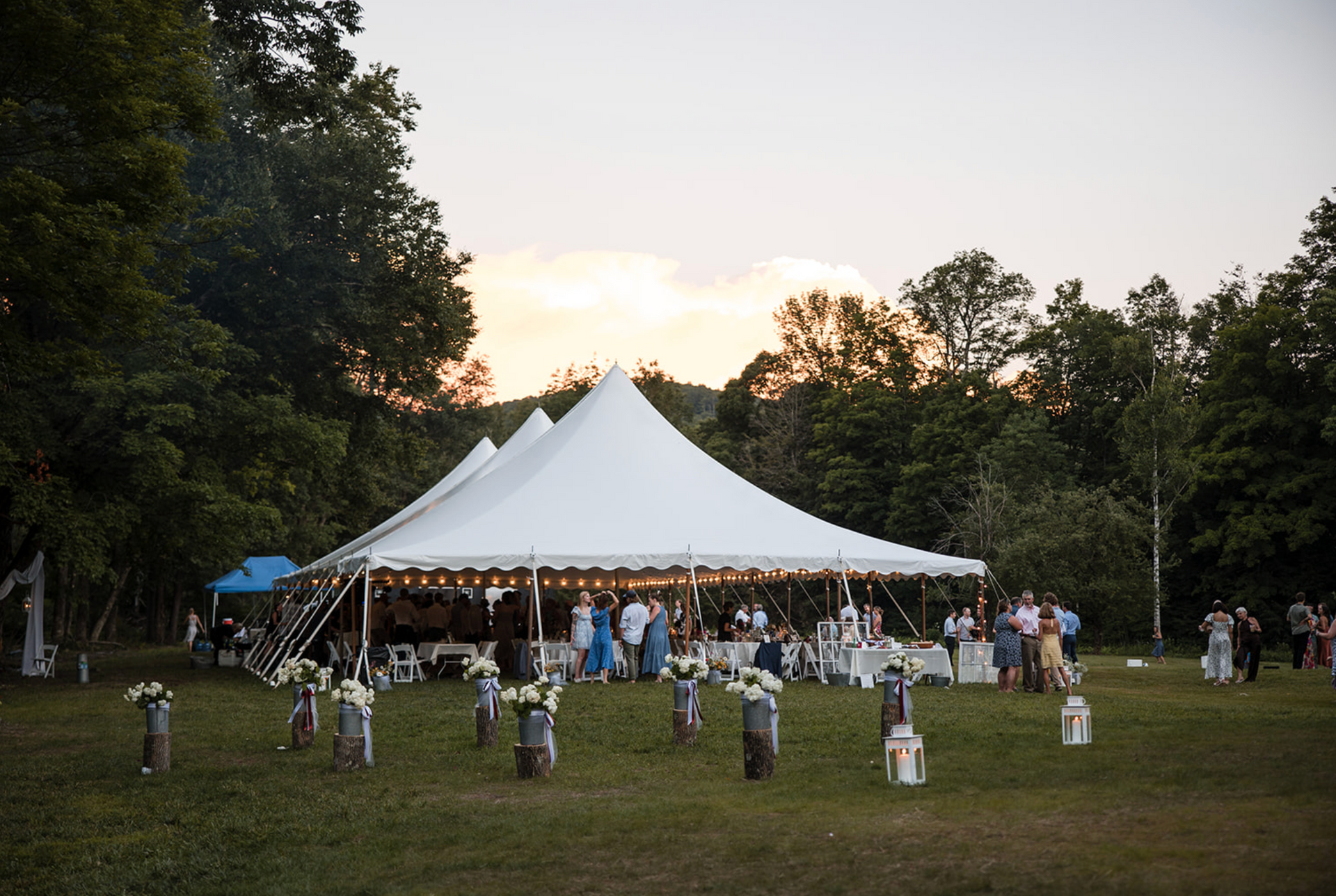 Wedding reception under a white tent in a grassy field; guests dine, and floral decorations are present.