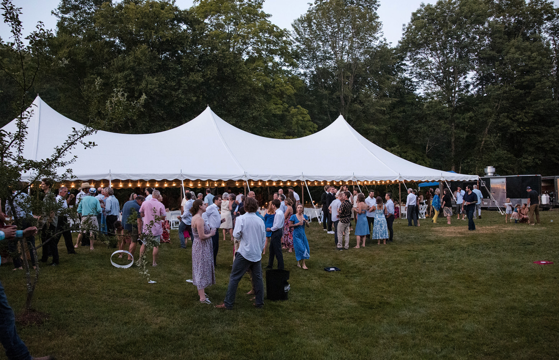People mingling on a grassy lawn at a white tented outdoor event. String lights are above.