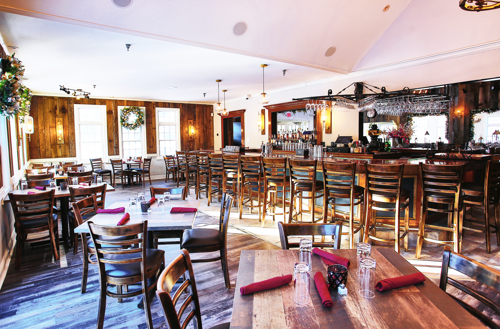 Restaurant interior: wooden tables/chairs, bar with stools, wood paneling, bright lights, holiday wreath on the wall.