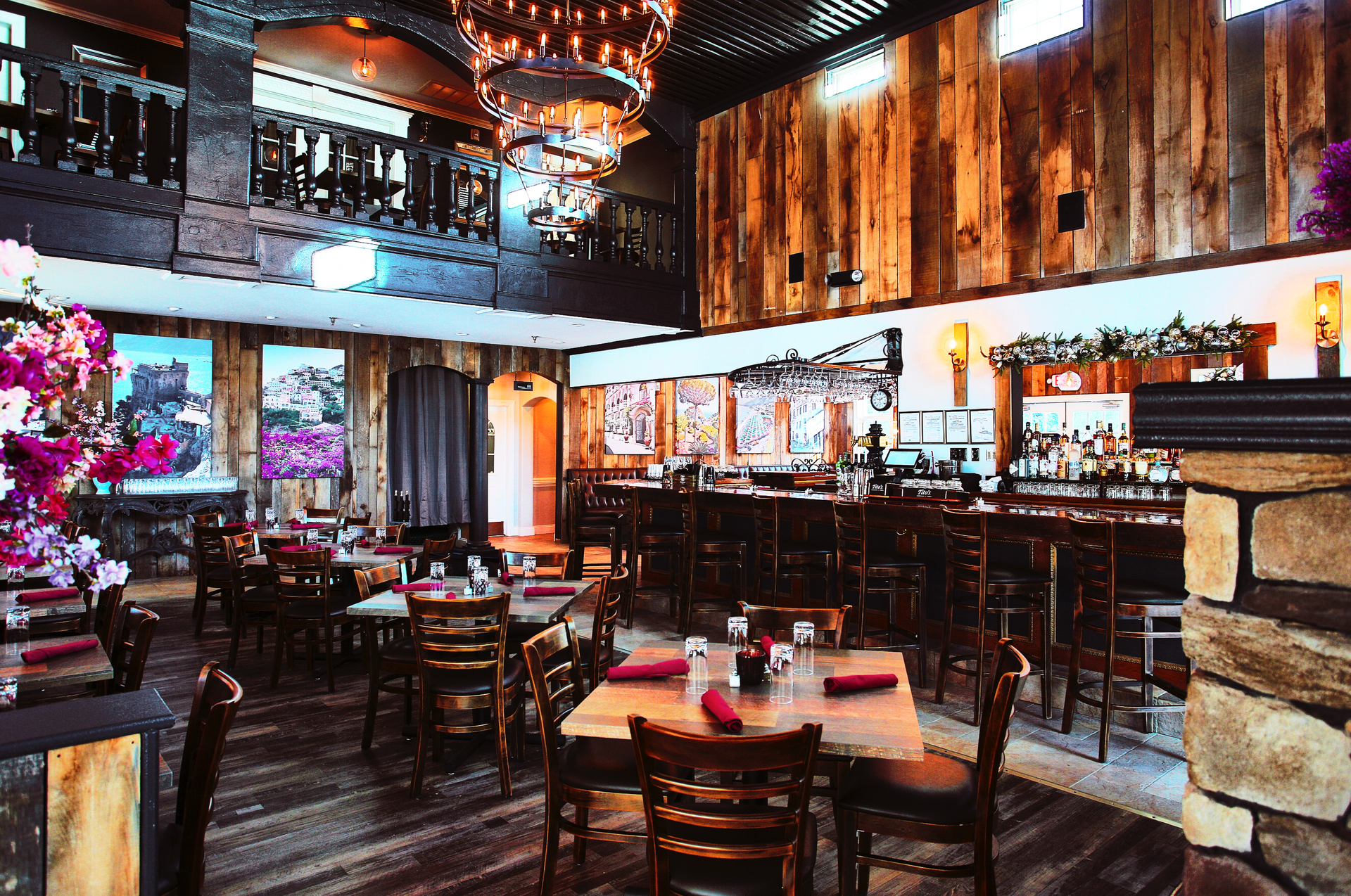 Interior of a rustic restaurant: wooden walls, bar, tables with red napkins, and a second-floor balcony.