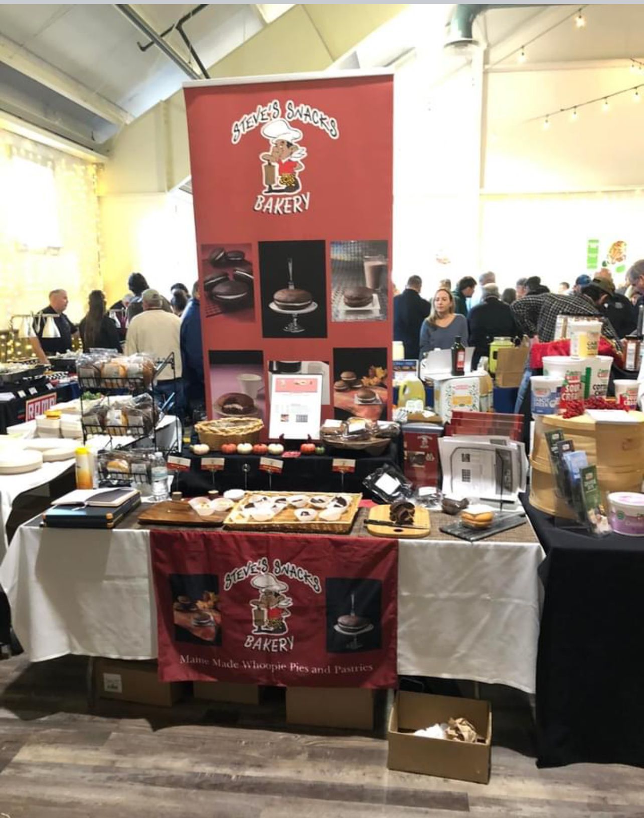 Bakery booth with red banner at an event, displaying baked goods like cakes and brownies. People browse nearby.