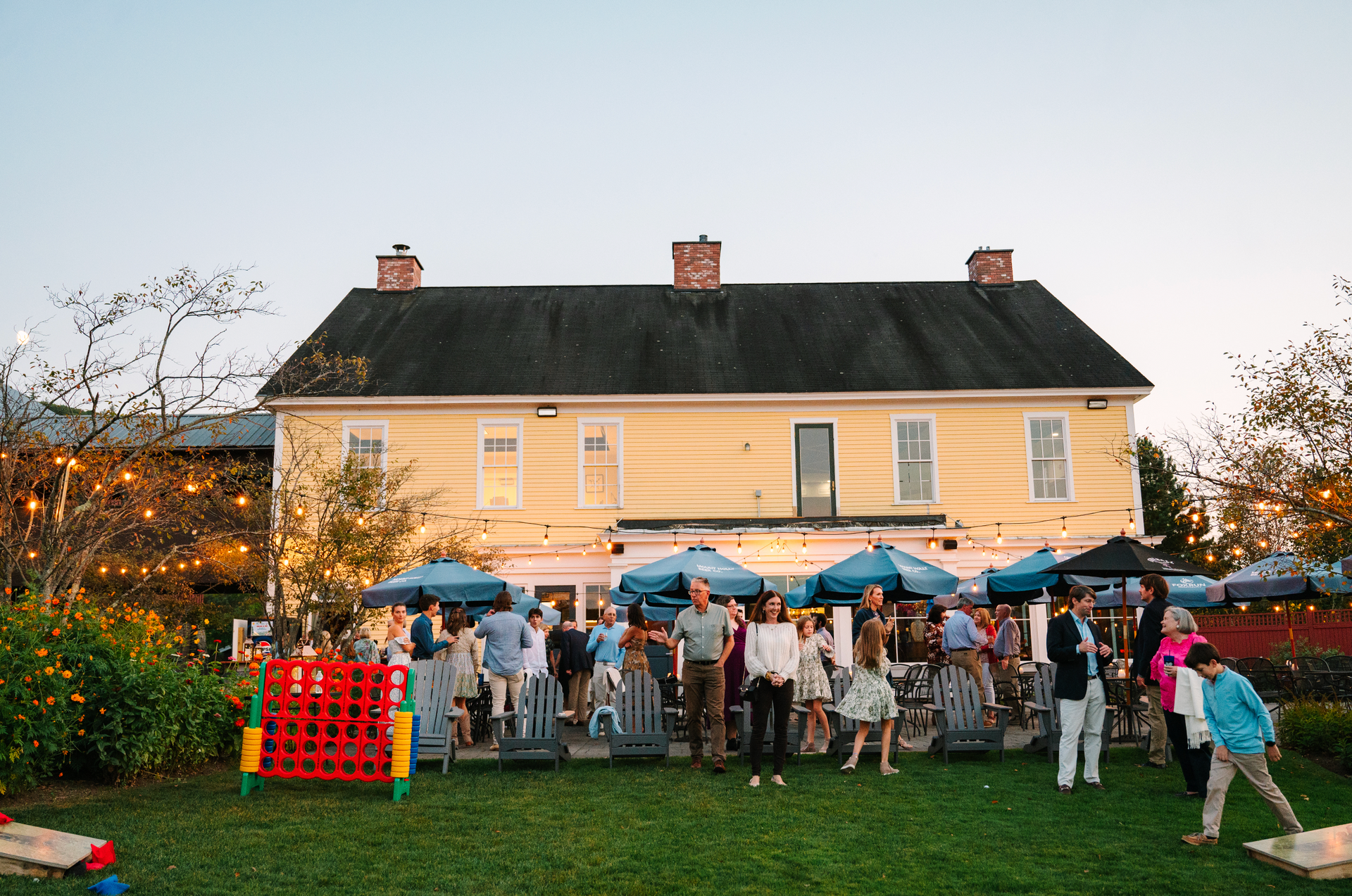 People gather on a lawn in front of a yellow building, some with umbrellas, possibly at an outdoor event.