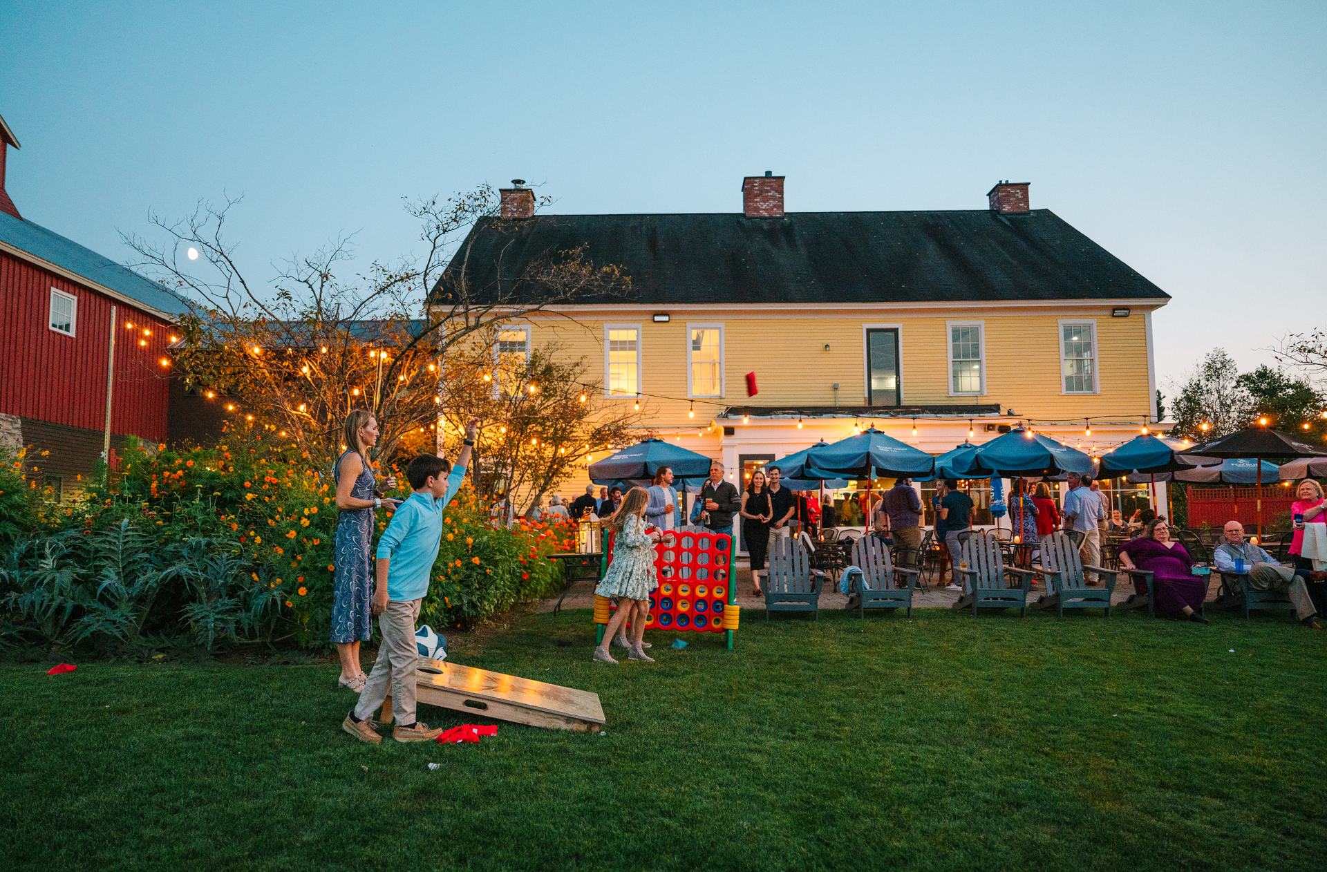 People playing cornhole on a lawn at dusk, with a yellow house and string lights in the background.