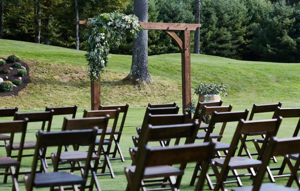A row of wooden folding chairs are lined up on a lush green field.