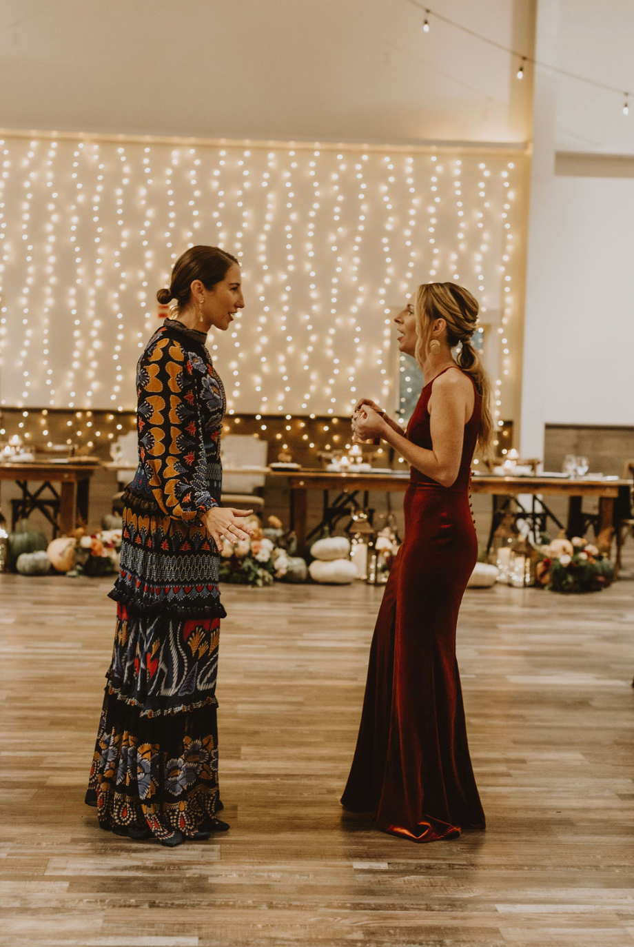 Two women in long dresses are standing next to each other on a wooden floor.
