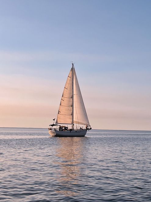 Sailboat on water, with a large white sail against a pastel sky.