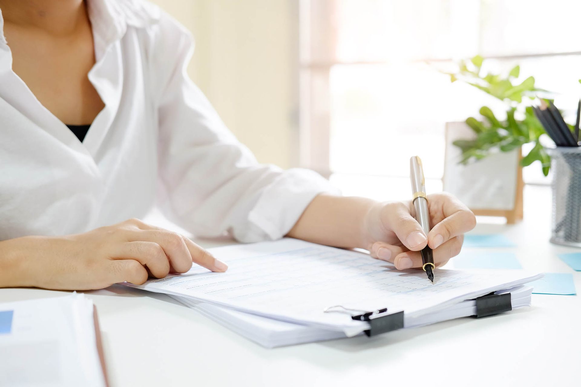 Person writing on a clipboard at a bright desk with a white shirt