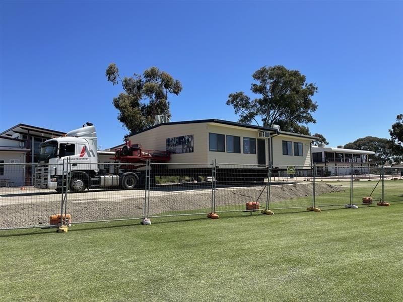 Mobile home being transported on a flatbed truck beside a fenced grassy lot under a clear blue sky