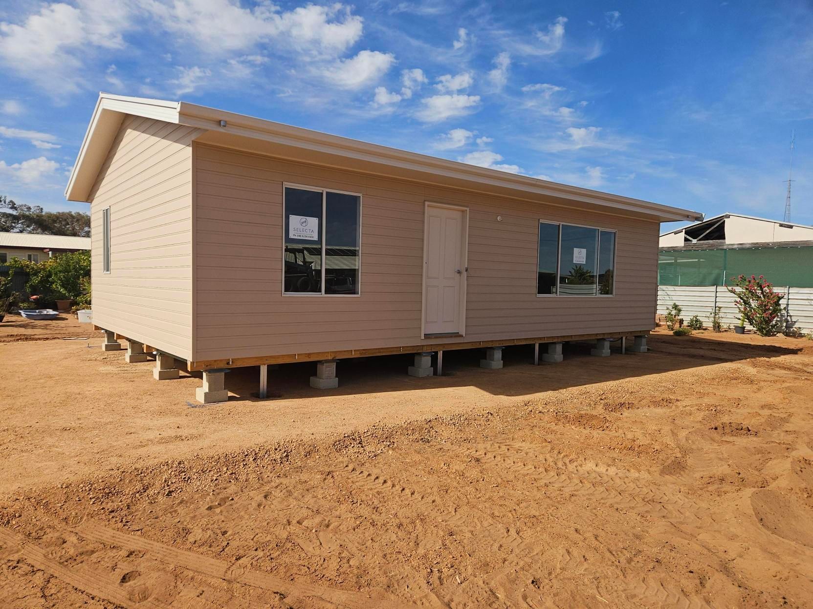 Small beige house under construction on a dirt lot, with windows and a slanted roof under a blue sky.