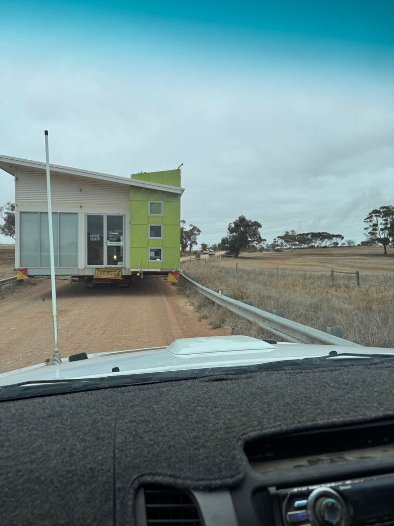 Small green tiny house being towed on a dirt road in a rural field, seen from inside a car. Small green tiny house being towed on a dirt road in a rural field, seen from inside a car.