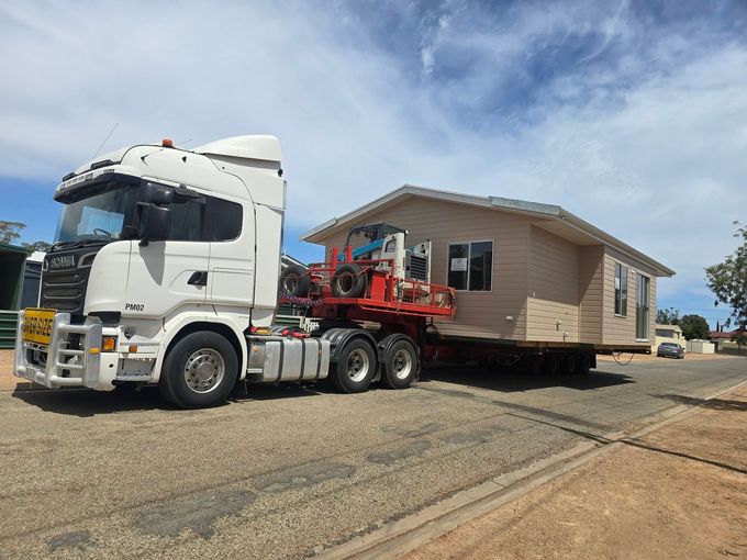White semi-truck hauling a beige house on a flatbed trailer along a roadside under a blue sky White semi-truck hauling a beige house on a flatbed trailer along a roadside under a blue sky