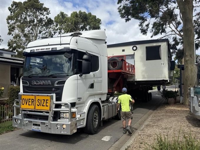 Large white truck carrying an oversized modular house on a residential street, with a worker guiding it. Large white truck carrying an oversized modular house on a residential street, with a worker guiding it.
