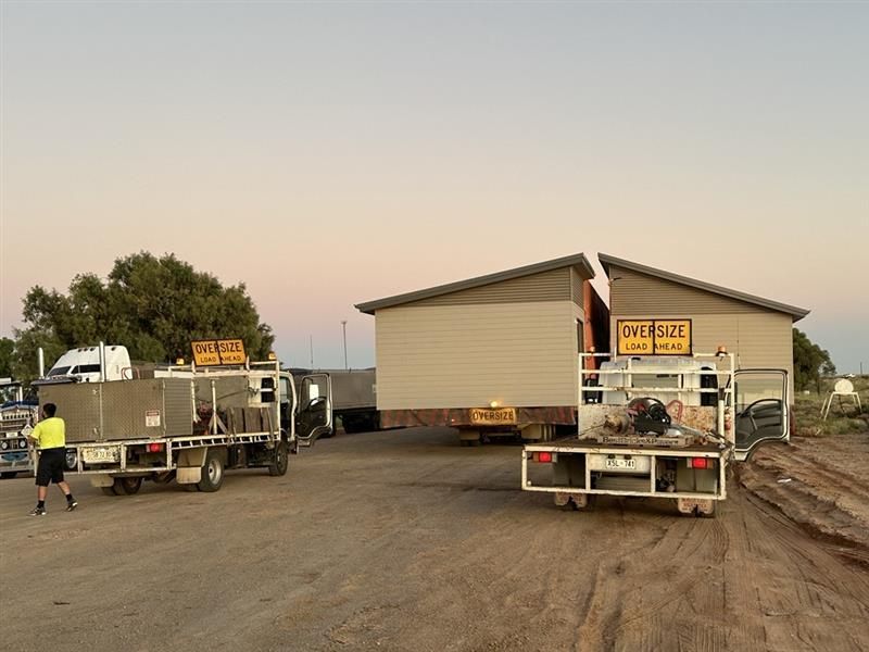 House being moved on a flatbed trailer at dusk, with trucks and workers on a dirt road. House being moved on a flatbed trailer at dusk, with trucks and workers on a dirt road.