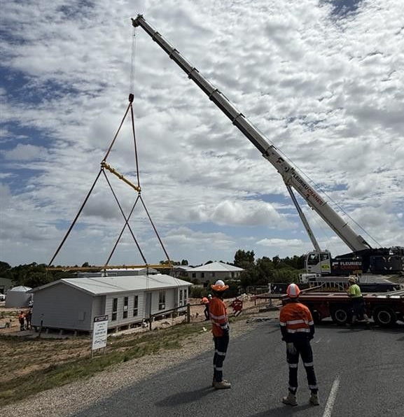 Gray shed on a trailer in a dusty open lot, with a tree and blue sky in the background Workers guide a crane lifting a large structure beside a road under a cloudy sky.