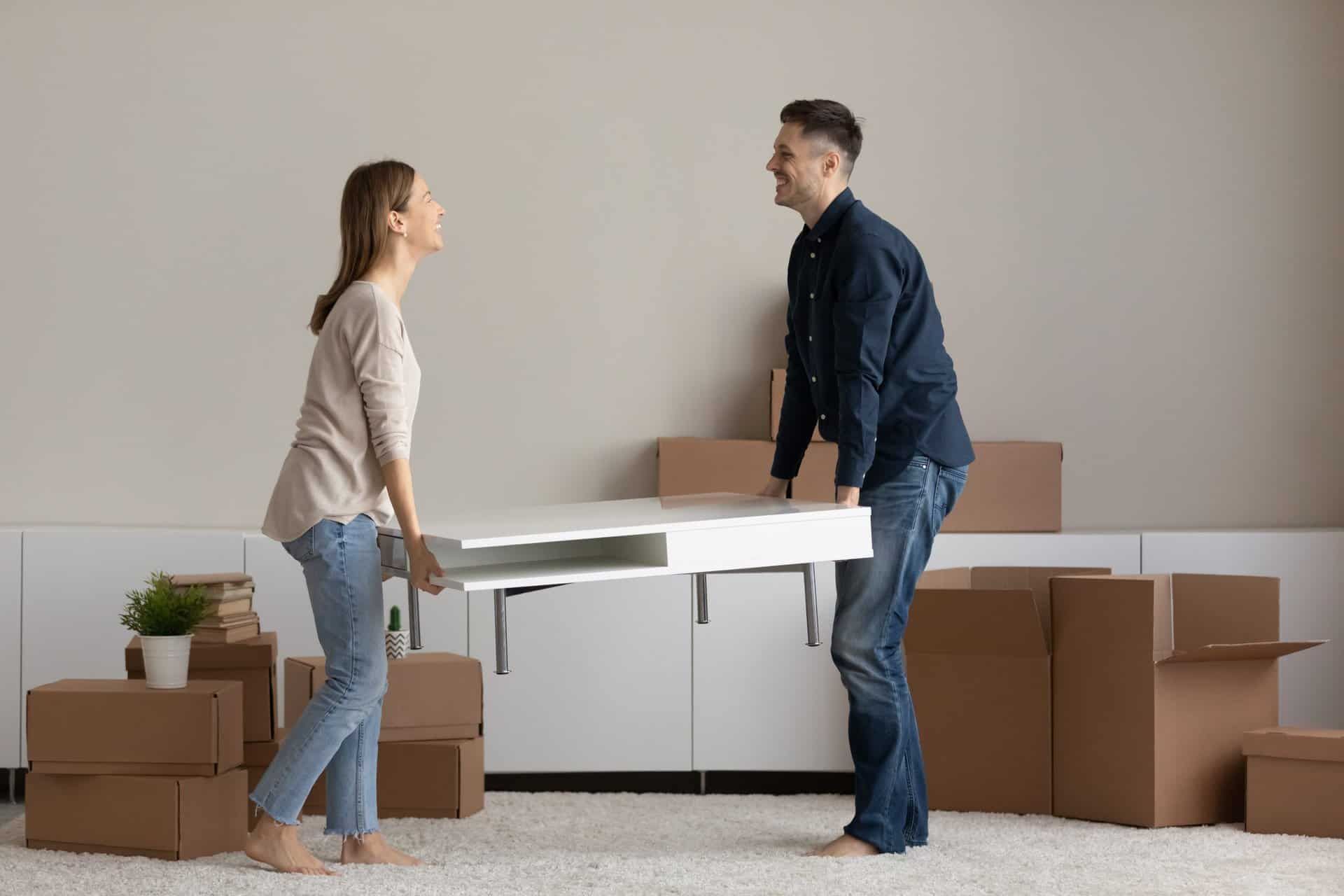 Happy millennial couple carrying coffee table together to unfurnished living room furnish new house