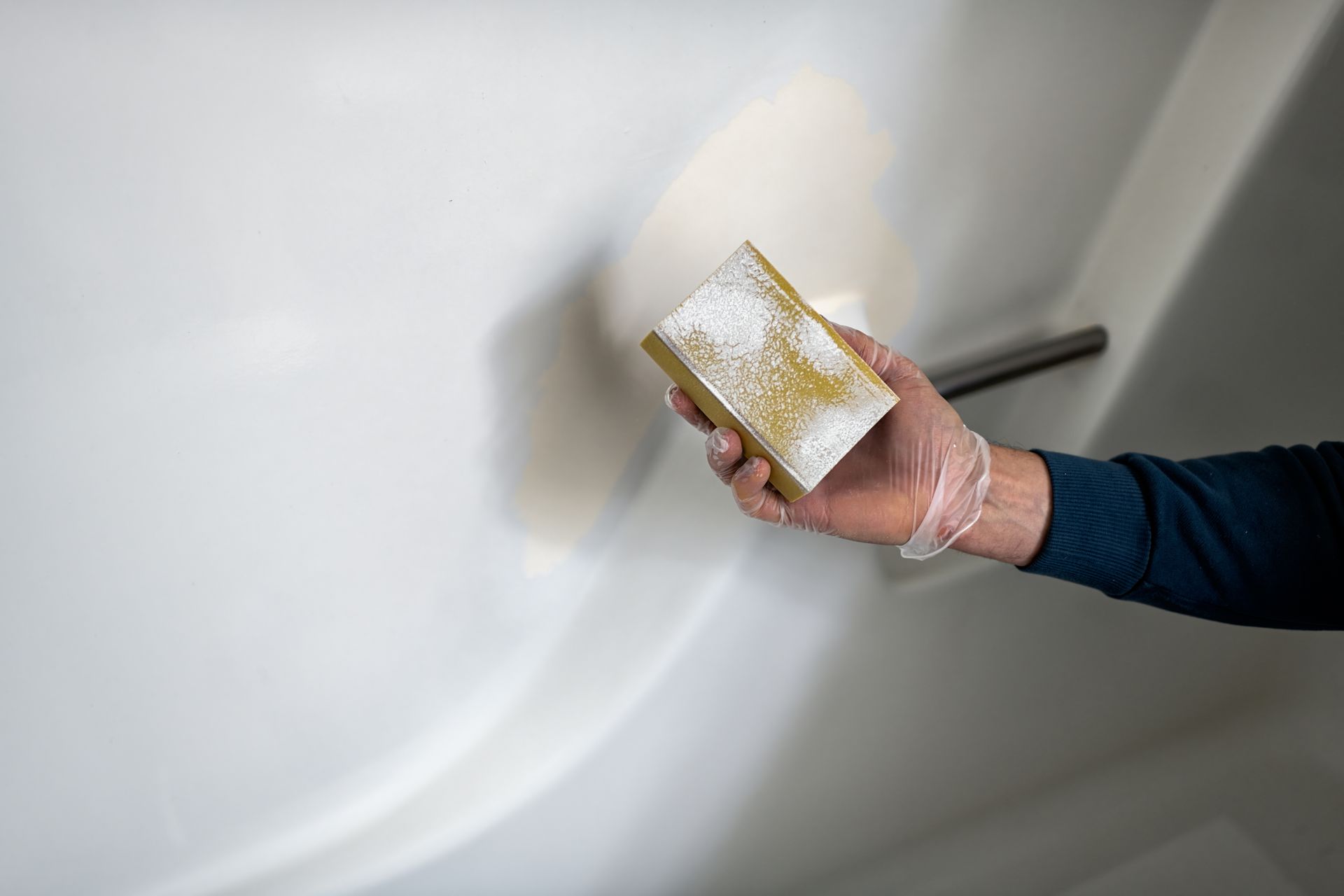 A hand holds a sanding block next to a painted and sanded bathtub wall.