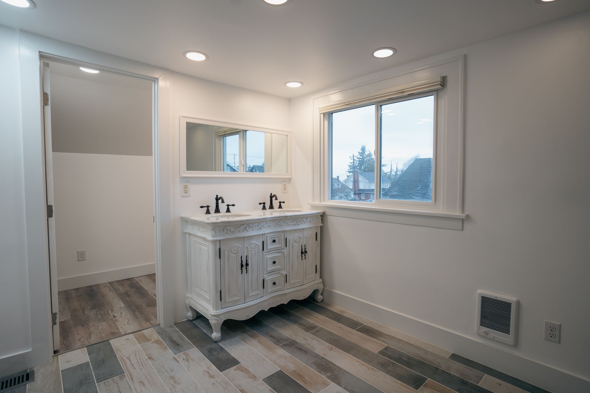 Bathroom with white vanity, double sinks, mirror, window, and wood-look flooring.