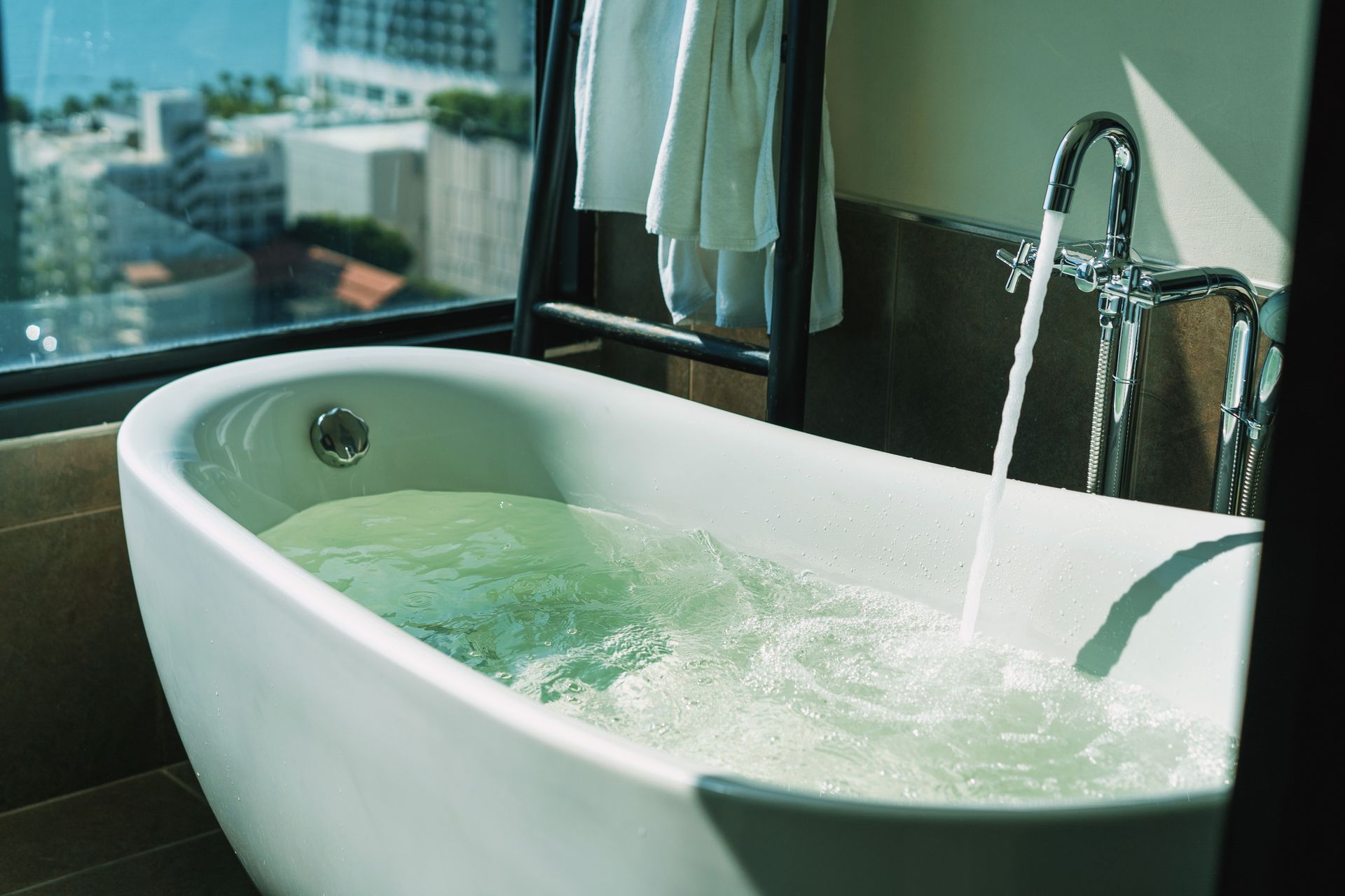 A bathtub fills with water, near a window with a city view. A towel hangs nearby.