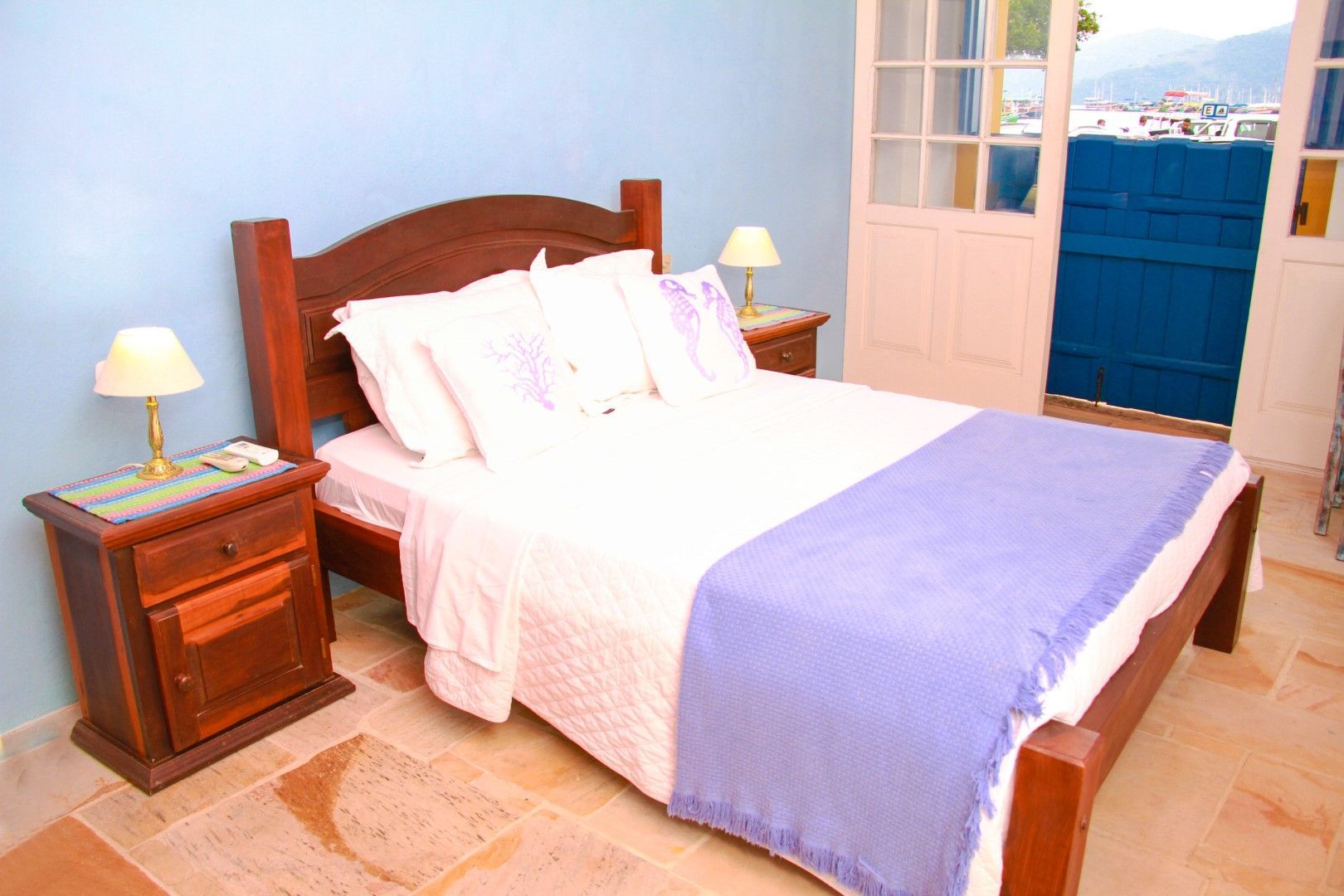 Um quarto com uma cama de madeira, dois criados-mudos, paredes azuis e uma porta aberta para uma varanda com vista para a cidade.