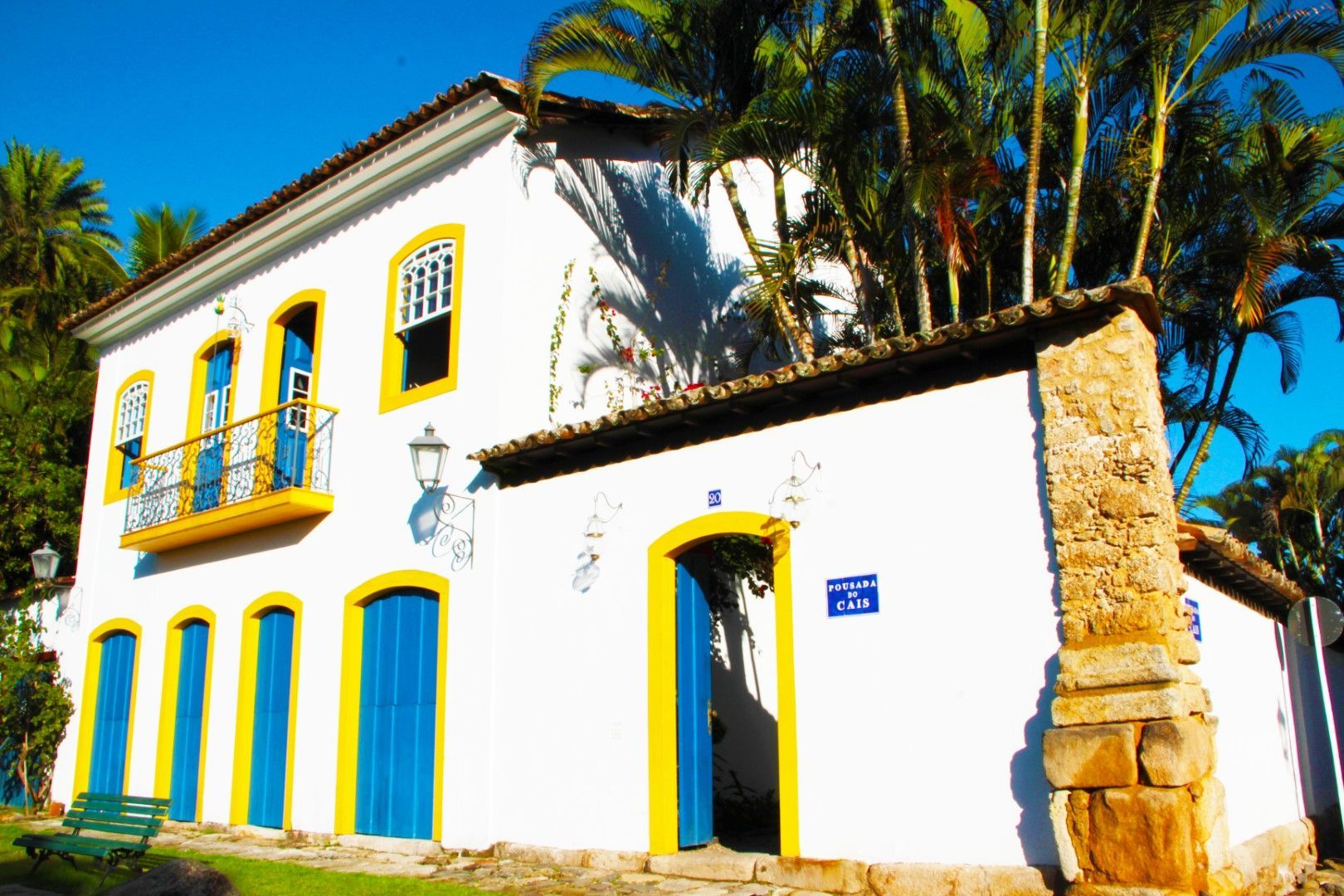 Edifício branco em estilo colonial com portas azuis e detalhes amarelos em um ambiente tropical. Palmeiras e um banco são visíveis.