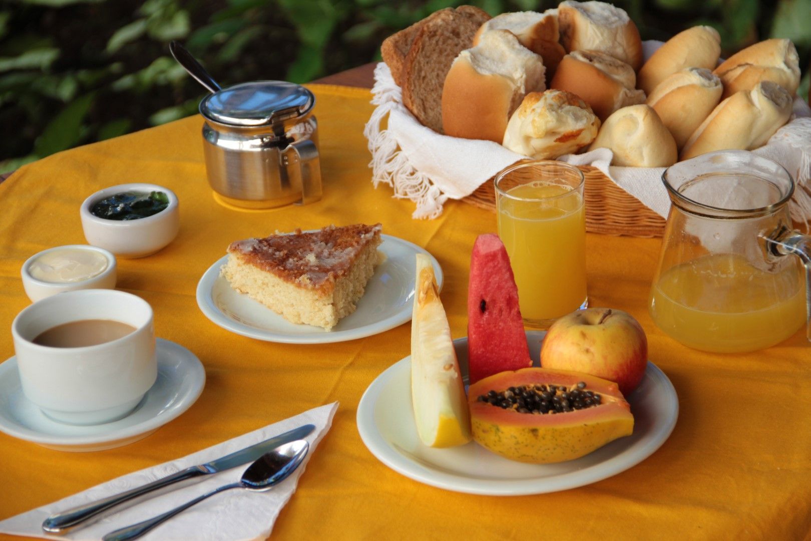 Café da manhã com pão, frutas, bolo, café, suco e condimentos em uma toalha de mesa amarela.
