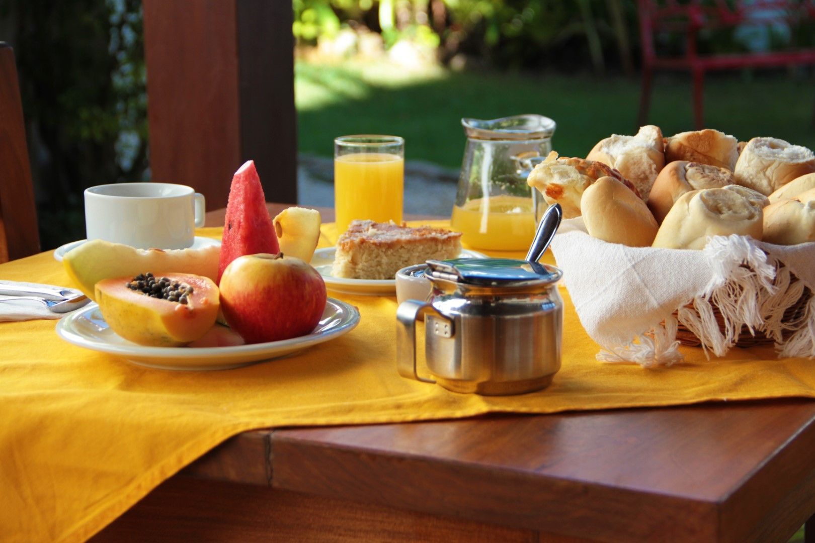 Mesa de café da manhã ao ar livre com frutas, pão, suco, café e bolo sobre uma toalha amarela. A luz do sol destaca a comida e o cenário.
