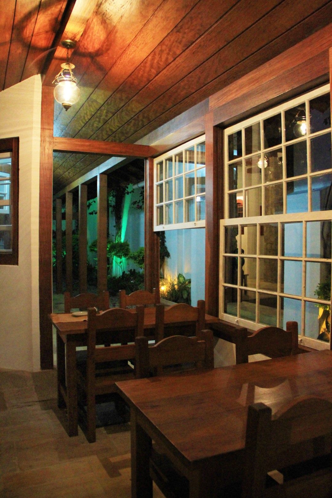 Mesas e cadeiras de madeira em uma área de jantar aconchegante e rústica, com grandes janelas com vista para um espaço externo iluminado.